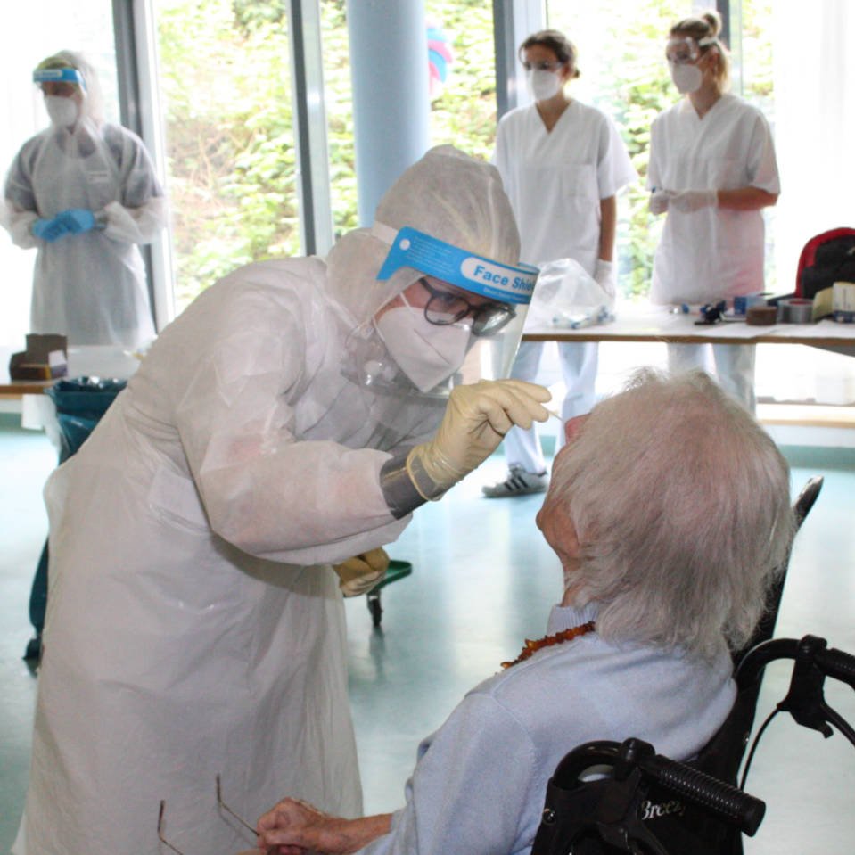 Foto einer Seniorin im Rollstuhl, bei der eine Hygienefachkraft in Schutzkleidung einen Nasen-Rachen-Abstrich vornimmt.